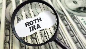 A magnifying glass over a stack of money with Roth IRA written over it. Signifying the amount of money to be saved with a Roth IRA account.