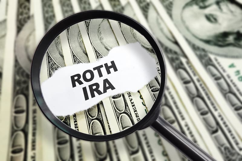 A magnifying glass over a stack of money with Roth IRA written over it. Signifying the amount of money to be saved with a Roth IRA account.