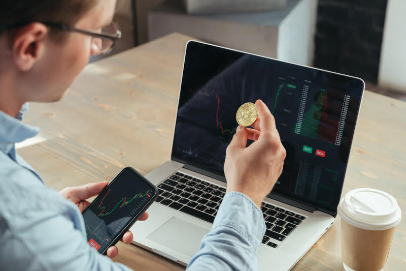 A man holding a gold coin in front of a laptop, comparing gold and bitcoin investment