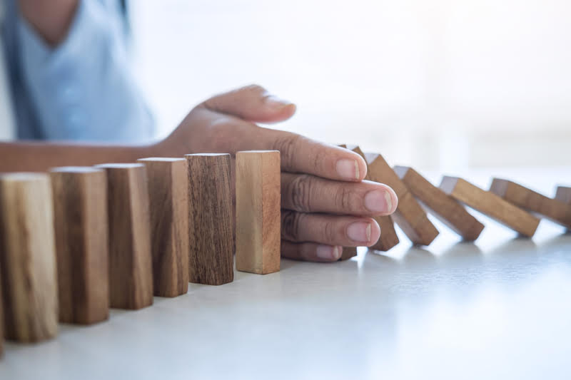 A person sitting at a table stopping wooden dominos from falling over, symbolizing strategically managing risk.